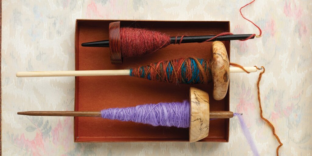 This image shows three different drop spindles resting in a red box. The spindles each have yarn wrapped around them (red, blue, and purple).