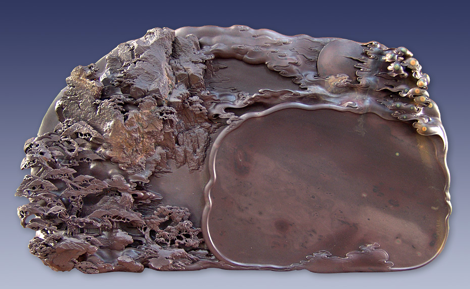 This image shows an inkstone carved with a nature scene of trees and mountains. The stone itself is a brown color.